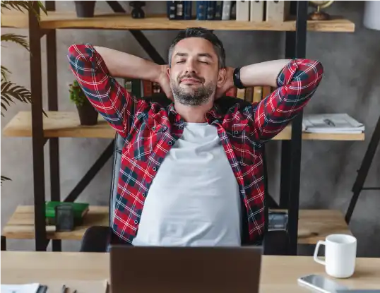 Relaxed man in a red plaid shirt leaning back with hands behind his head, sitting at a desk with a laptop, looking satisfied.